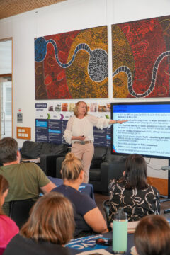 A woman stands in front of a seated audience, pointing to a presentation slide on a screen. Behind her are colorful, abstract wall artworks. The setting appears to be an educational or community event.
