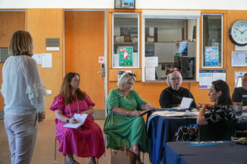 A group of five women sit and talk around tables in a brightly lit room with wooden doors, posters on the wall, and a clock showing 10:24. One woman stands while the others listen and take notes.