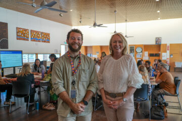 Two people stand smiling in the foreground of a brightly lit room where groups of people are seated at tables in conversation. The setting appears to be a casual community or workshop event.