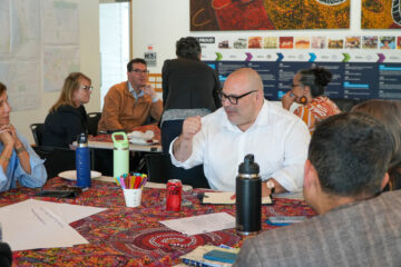 A group of people sit around a colorful table, engaged in discussion. One man in a white shirt gestures while others listen attentively. Papers, drinks, and pens are spread across the table in a bright, modern room.
