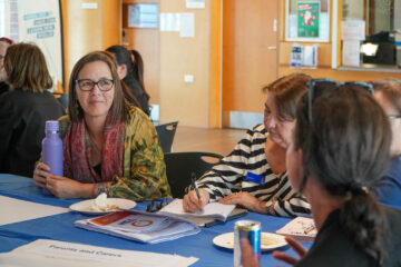 A group of adults sit around a table having a discussion. One woman holds a purple water bottle and listens attentively, while another writes notes. Papers and drinks are scattered on the table.