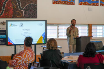 A man stands at a podium giving a presentation to a seated audience. A screen beside him displays a thank-you slide. Colorful artwork decorates the walls of the bright room.