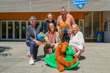 Five smiling adults kneel and squat outside a building, holding toys including a globe, abacus, and rocking horse with a stuffed lion. A youth center sign is visible above them.