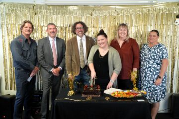 Six people stand in front of gold streamers; the woman in the center is cutting a chocolate cake on a table with fruit and decorations. They are all dressed in semi-formal attire and smiling at the camera.