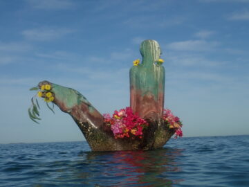 A colorful sculpture of a person partially submerged in the sea is decorated with flowers around its neck, leg, and base. The sky is clear and blue above the calm water.
