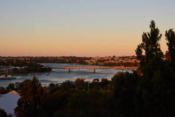 A river with boats passes under a low bridge at sunset, with houses and greenery on both banks and silhouetted trees in the foreground.