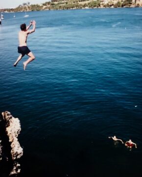 A person is mid-air, jumping off a rocky cliff into a large body of blue water, while two swimmers float below near the cliff. Trees and shoreline are visible in the background.