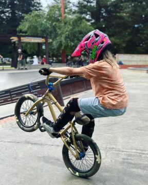 A person wearing a pink helmet, orange shirt, and protective gear performs a wheelie on a BMX bike at a skate park, with trees and other people in the background.