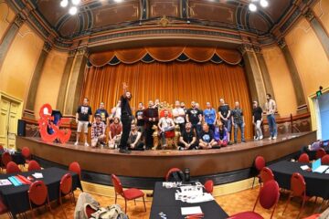 A group of people pose together on a stage in an ornate hall with red curtains, high ceilings, and decorative walls; tables and chairs are set up in the foreground.