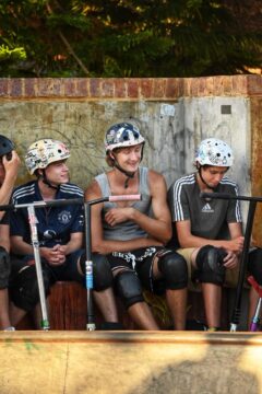 Three young people wearing helmets and knee pads sit with scooters at a skatepark. The person in the center is smiling and pointing, while the others watch and listen. A brick wall and greenery are in the background.