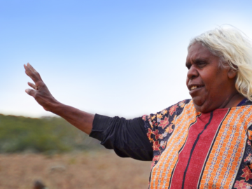 An older woman with long white hair, wearing a colorful patterned top, raises her hand in greeting or gesture against a clear blue sky and natural landscape.
