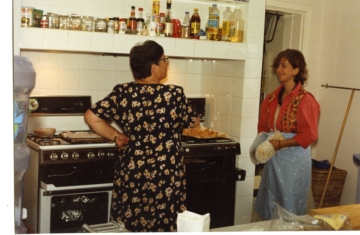 Two women are in a kitchen. One, wearing a floral dress, cooks at the stove while the other, in a red shirt and apron, stands nearby holding an oven mitt. Shelves of spices and condiments are visible above the stove.