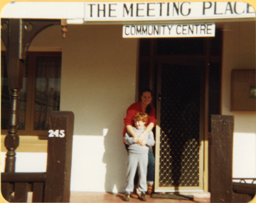 A woman and a child stand smiling in front of the entrance to a building labeled The Meeting Place Community Centre with the number 245 visible on the porch post.
