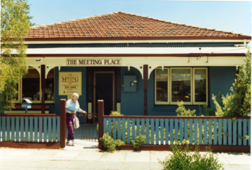 An elderly woman opens a gate in front of a blue house with a red roof and sign reading The Meeting Place. The house is surrounded by a fence and greenery, and the sun is shining.