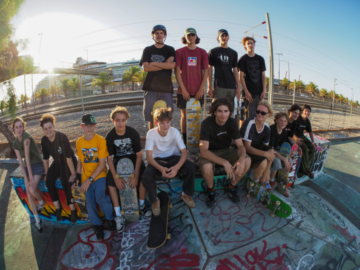 A group of skateboarders, teens and young adults, pose together at a graffiti-covered skate park on a sunny day, holding their skateboards and smiling at the camera.