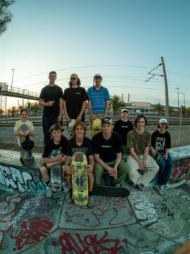 A group of eleven skateboarders pose together on a graffiti-covered ledge at a skatepark, holding skateboards, with train tracks, palm trees, and an industrial background under a clear sky.