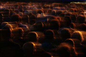 A large crowd of people is seen from behind, illuminated by warm, low lighting with blurred motion effects, creating an atmospheric and energetic scene.