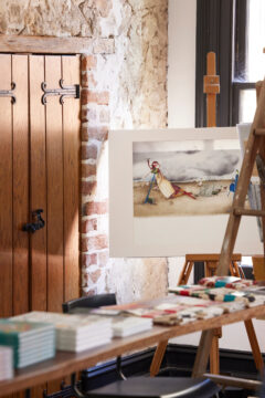 An art studio with a watercolor painting on an easel, depicting people and a kite on a windy day. The room has rustic stone walls, wooden doors, stacked books, and various art supplies on a table in the foreground.
