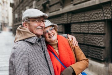 An elderly couple, both smiling, stand close together on a city street. The man wears a gray cap and plaid coat, and the woman wears glasses, a knit hat, a bright red scarf, and a tan coat.