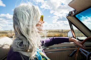 A woman with long, silver hair and sunglasses drives a convertible car with the top down on a sunny day, wind blowing her hair, and an open rural landscape in the background.