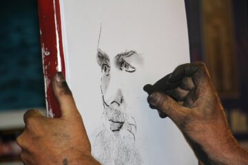 A close-up of an artist’s hands sketching a realistic face on white paper with charcoal; the artist’s hands and fingers are covered in dark charcoal smudges.