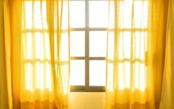 A sunlit window with sheer yellow curtains partially drawn, allowing bright sunlight to stream into the room and create a warm, glowing atmosphere.