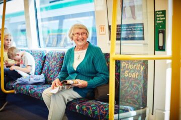 An elderly woman with glasses and a teal sweater sits and smiles on a train’s priority seat, holding a newspaper. Other passengers, including a young boy, are seated nearby. A green sign and a window are visible.