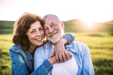 An older couple smiling and embracing outdoors in a sunlit field, both wearing casual denim shirts, with green hills and a bright sun in the background.