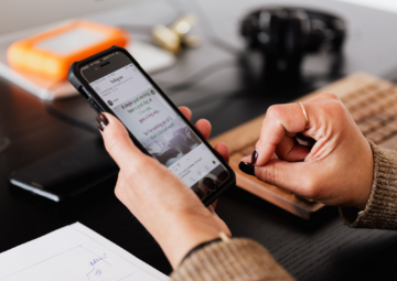 A person holding a smartphone and scrolling through an Instagram feed at a desk, with a keyboard, headphones, and documents nearby.