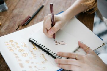 A person practices calligraphy with a brown pen in a spiral notebook, writing the word love. Sheets with cursive writing examples and pens are scattered on the wooden table.