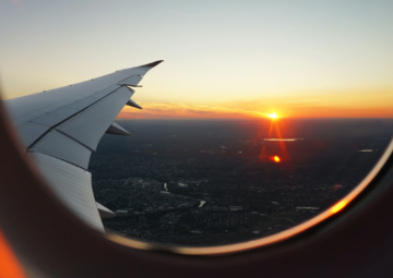View from an airplane window showing the wing and a vibrant sunset over a landscape, with the sun low on the horizon and an orange glow spreading across the sky.