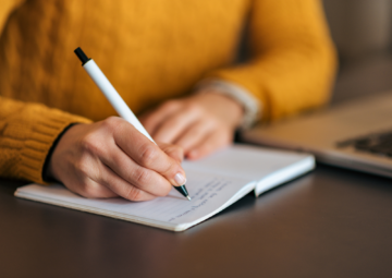 A person wearing a yellow sweater writes in a notebook with a white pen, seated at a table next to a laptop. The focus is on the hand and notebook, with the background softly blurred.