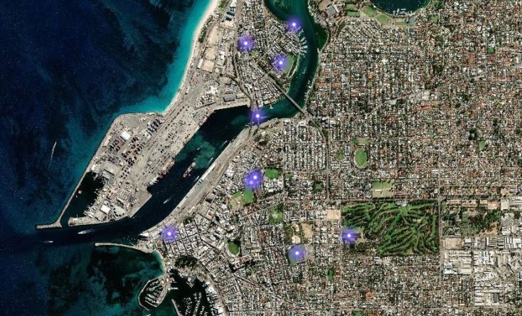 A satellite view of a coastal city showing a harbor, urban areas, green parks, and several blue markers indicating specific locations throughout the city.