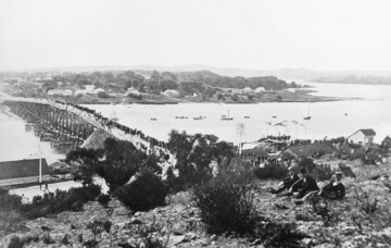 Black-and-white photo of a long wooden bridge crowded with people, crossing a river toward a small town. In the foreground, three people sit on a hill, overlooking the scene. Boats are visible on the water below.