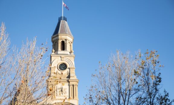 A tall clock tower with a pointed roof and a flag on top stands against a clear blue sky, partially framed by leafless trees.