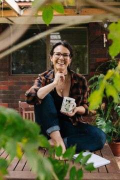 A smiling woman wearing glasses and a plaid shirt sits cross-legged on an outdoor wooden table, holding a mug and a pen, with greenery and a red brick house in the background.