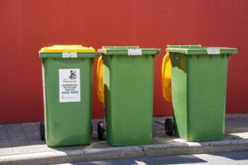 Three green wheelie bins with yellow lids are lined up on a sidewalk against a red wall. A white label with contact information is visible on the front of the leftmost bin.