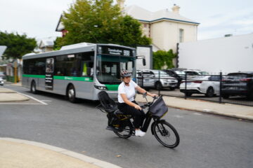 A person wearing a helmet rides an electric bike on a city street, while a public bus passes by in the background. Parked cars and residential buildings line the street.