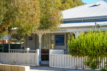 A white building with a tin roof and a veranda, partially shaded by trees. A sign reads The Meeting Place. A white picket fence and green shrubs are in front. Sunlight filters through the foliage.