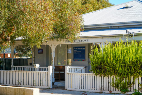 A white building with a tin roof and a veranda, partially shaded by trees. A sign reads The Meeting Place. A white picket fence and green shrubs are in front. Sunlight filters through the foliage.