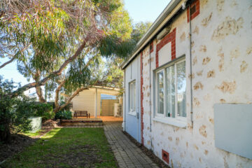 A rustic house with stone and brick walls features white-framed windows. A paved path runs alongside the house, leading to a wooden bench and covered patio area beneath large leafy trees.