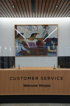 A customer service desk with microphones and clear dividers, featuring a sign that reads CUSTOMER SERVICE Welcome Wanjoo. A colorful painting of four people hangs on the wall behind the desk.
