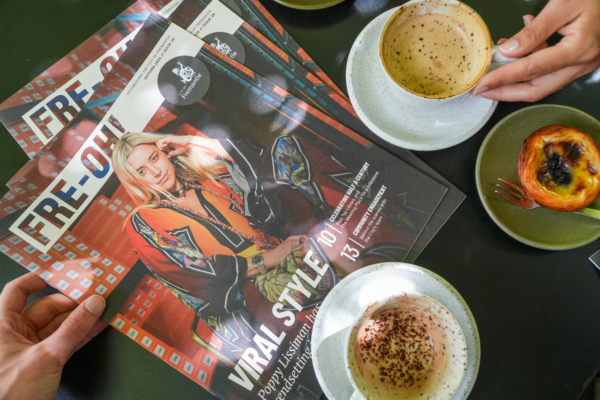 Two people sit at a table with coffee cups, a pastry, and two magazines featuring a blonde woman on the cover titled “VIRAL STYLE.” One hand holds a magazine, while the other rests near a cappuccino.