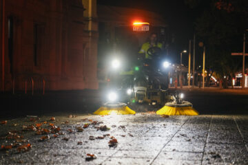 A street sweeper with bright lights cleans leaves and debris from a city sidewalk at night, with a worker visible behind the machine.