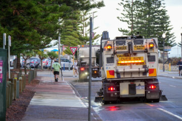 Two street cleaning trucks drive down a coastal road while a worker in a green shirt cleans the sidewalk. Tall pine trees line the street, and a few pedestrians walk nearby. The scene appears to be early morning or late afternoon.