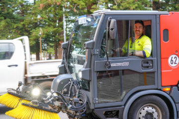 A man in a high-visibility shirt is smiling while operating a street cleaning vehicle with yellow brushes on a city road. Trees and a white truck are visible in the background.