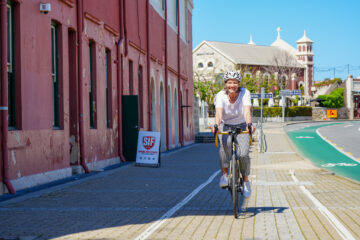 A woman wearing a helmet rides a bicycle on a sunny day along a sidewalk next to a red building, with a church and clear blue sky in the background.