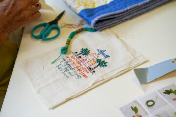A person embroiders a piece of fabric showing a house, trees, and text. Green thread, scissors, and a folded quilt are nearby on the white table.