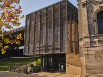 A modern glass building with a perforated metal facade and the words City of Fremantle Walyalup Civic Centre on the front, next to a historic stone building; a tree with yellow leaves stands to the left.