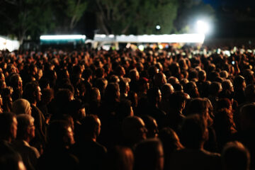 A large crowd of people gathered outdoors at night, facing towards bright lights in the distance, with trees and structures visible in the background.